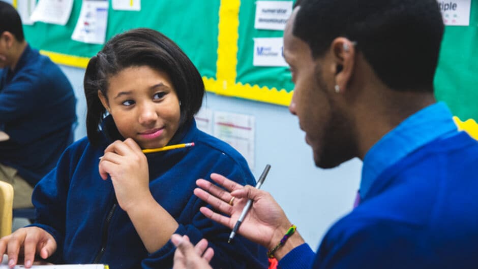 A female student with a pencil under her chin looking towards her teacher.