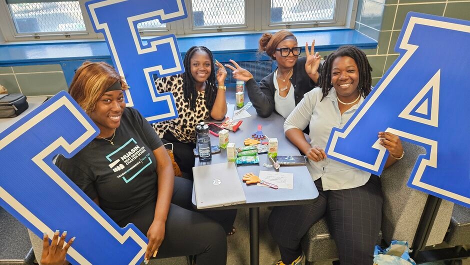 A group of corps members sit in a cafeteria holding blue TFA letters.