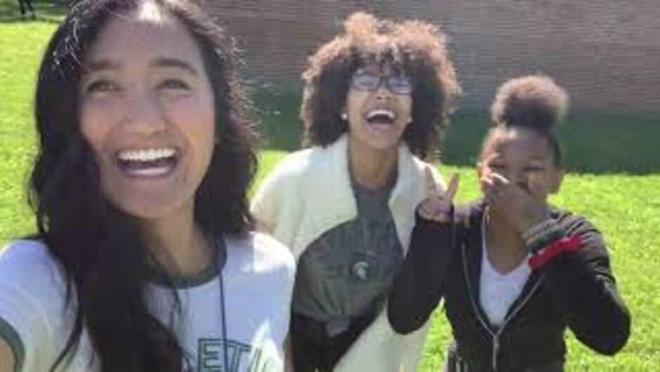 A woman smiles in the foreground with two others in the background.