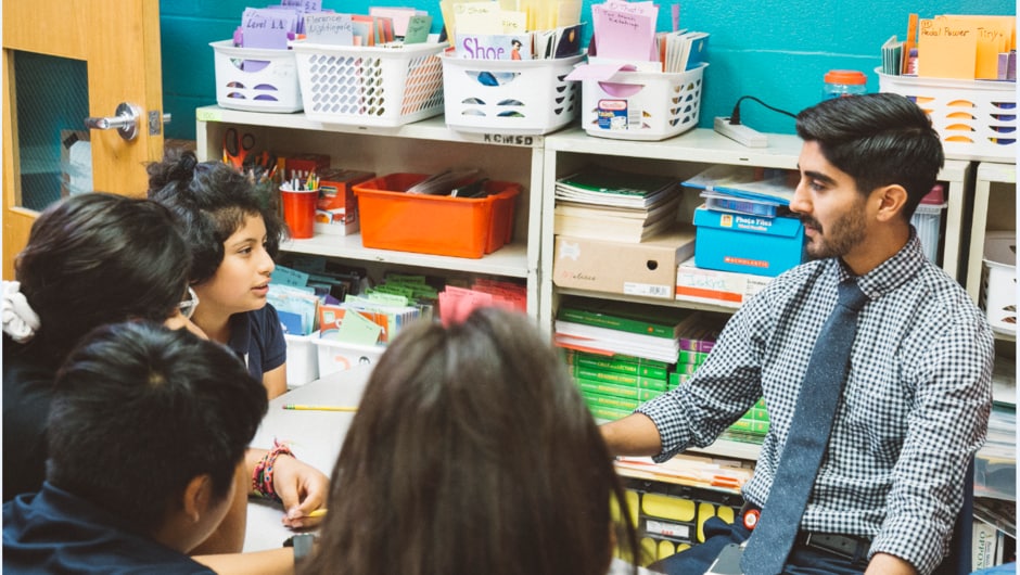 A teacher sits across a table from students. The classroom wall is bright blue.