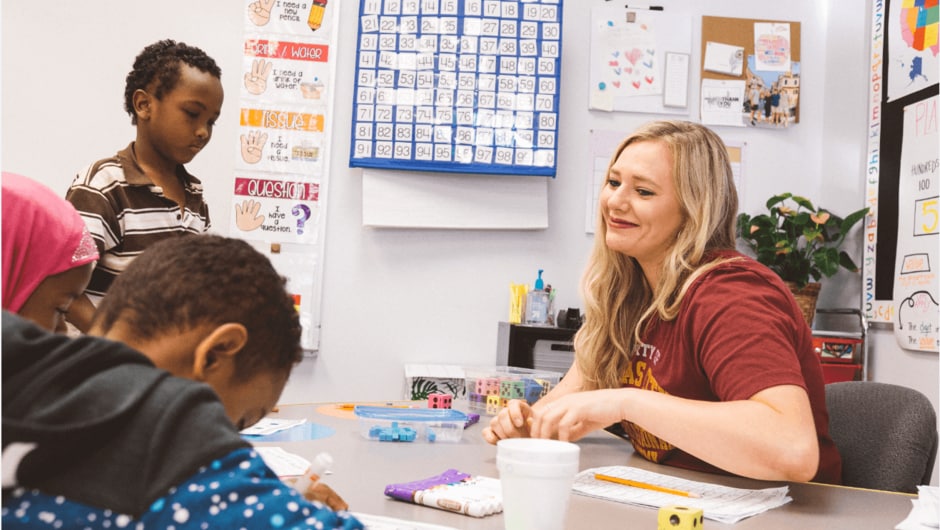 A teacher smiles across a table at young students