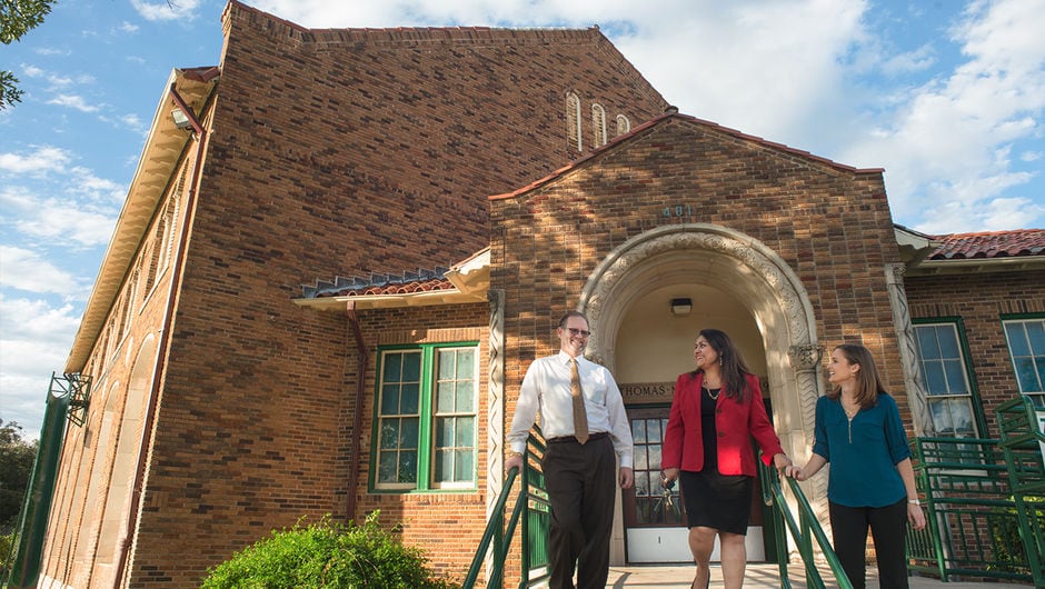 One middle-aged male and two young adult females walk down the stairs outside of a large brick school building.