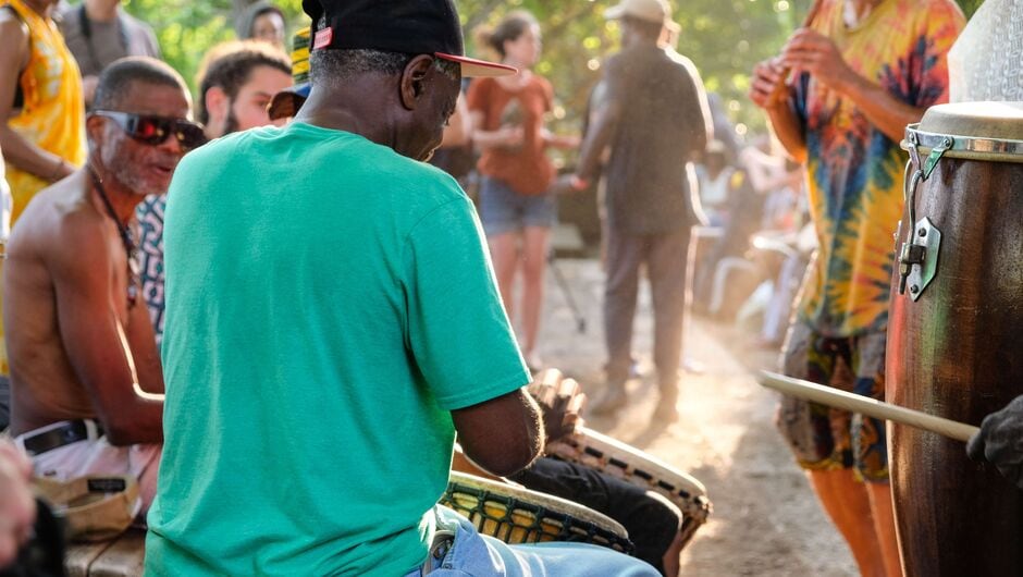 People drum in a circle as others listen, outdoors.